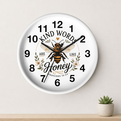 A round white wall clock with a printed bee, floral details, and the words 'KIND WORDS ARE LIKE Honey' on the face, shown hanging on a light wall above a small potted succulent.