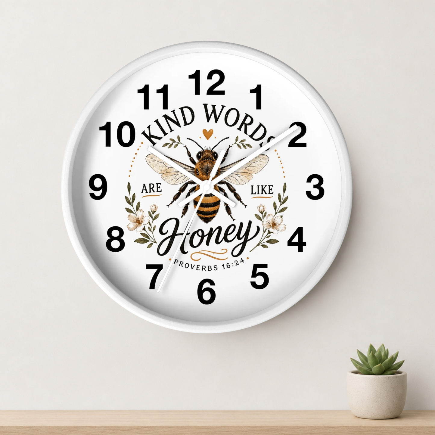 A round white wall clock with a printed bee and floral design, surrounded by the words 'KIND WORDS ARE LIKE Honey PROVERBS 16:24'. The clock is mounted on a light beige wall above a wooden shelf with a small potted succulent.