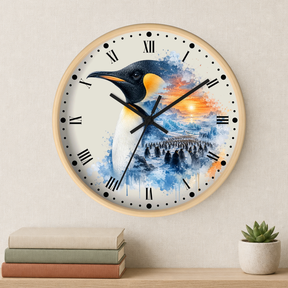 A round wall clock with a light wood frame hangs on a beige wall above a shelf with stacked books and a small potted succulent. The clock face features a watercolor-style penguin illustration over blue, white, and orange water/sky tones, with black Roman numerals and hands.