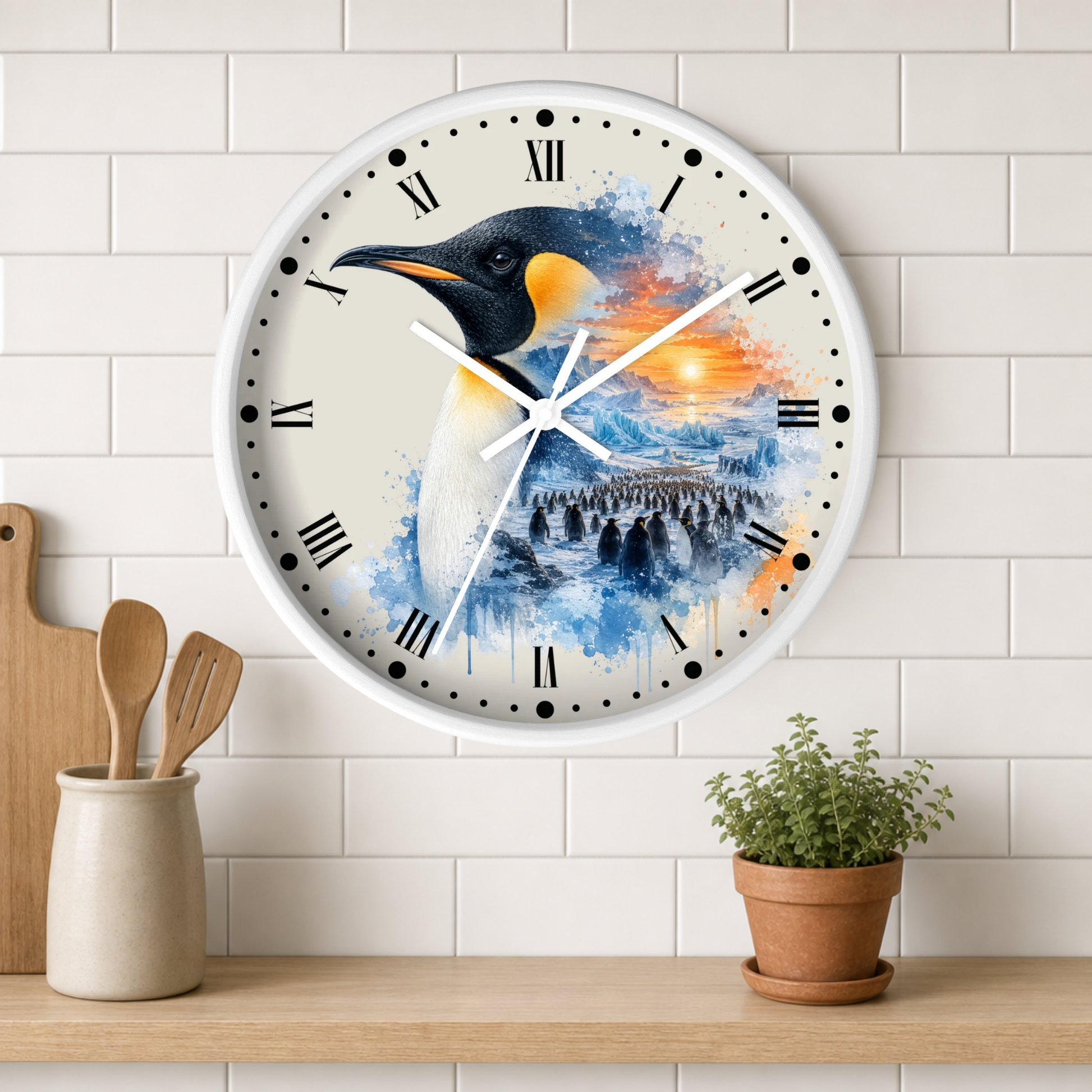 A round wall clock with a white frame and a penguin-themed watercolor design, mounted on a white subway tile wall above a wooden shelf with kitchen utensils and a potted plant.