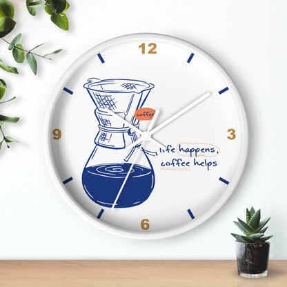 A round white wall clock with a coffee maker illustration and the phrase 'life happens, coffee helps' on the face. The clock is mounted on a white wall above a light wood surface, with green leaves and a small potted succulent nearby.