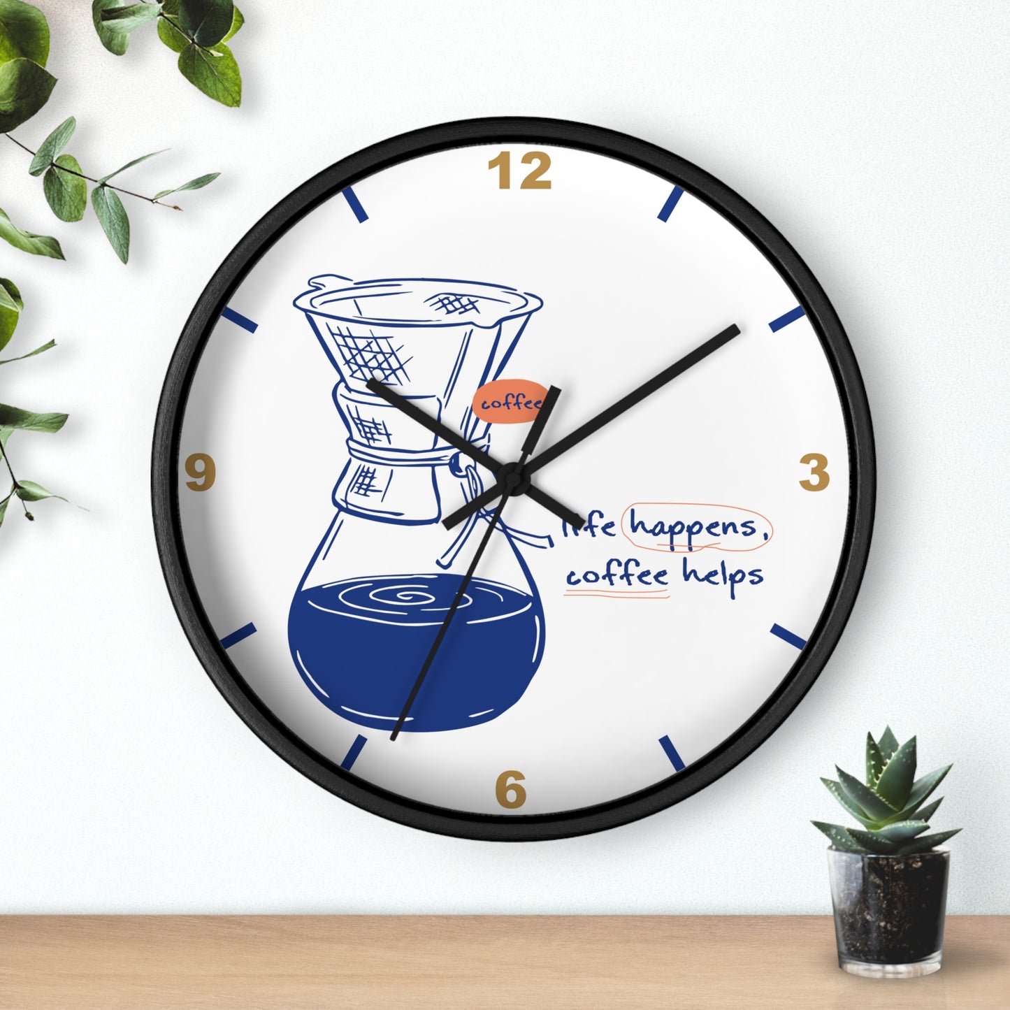 A round wall clock with a black frame and white face, featuring a coffee maker illustration, gold numbers, and the phrase 'life happens, coffee helps.' The clock is shown hanging on a white wall above a wooden surface with small green plants nearby.