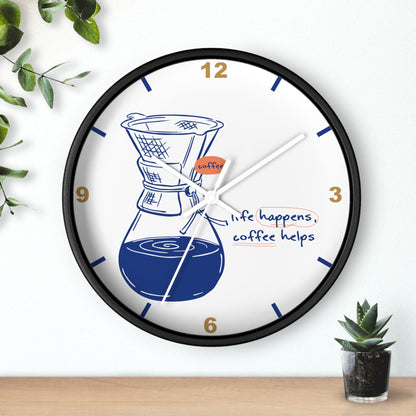 A round wall clock with a black frame and a white face, featuring a coffee maker illustration, gold numbers, and the phrase 'life happens, coffee helps.' The clock is shown hanging on a white wall above a wooden surface, with small green plants at the corners.