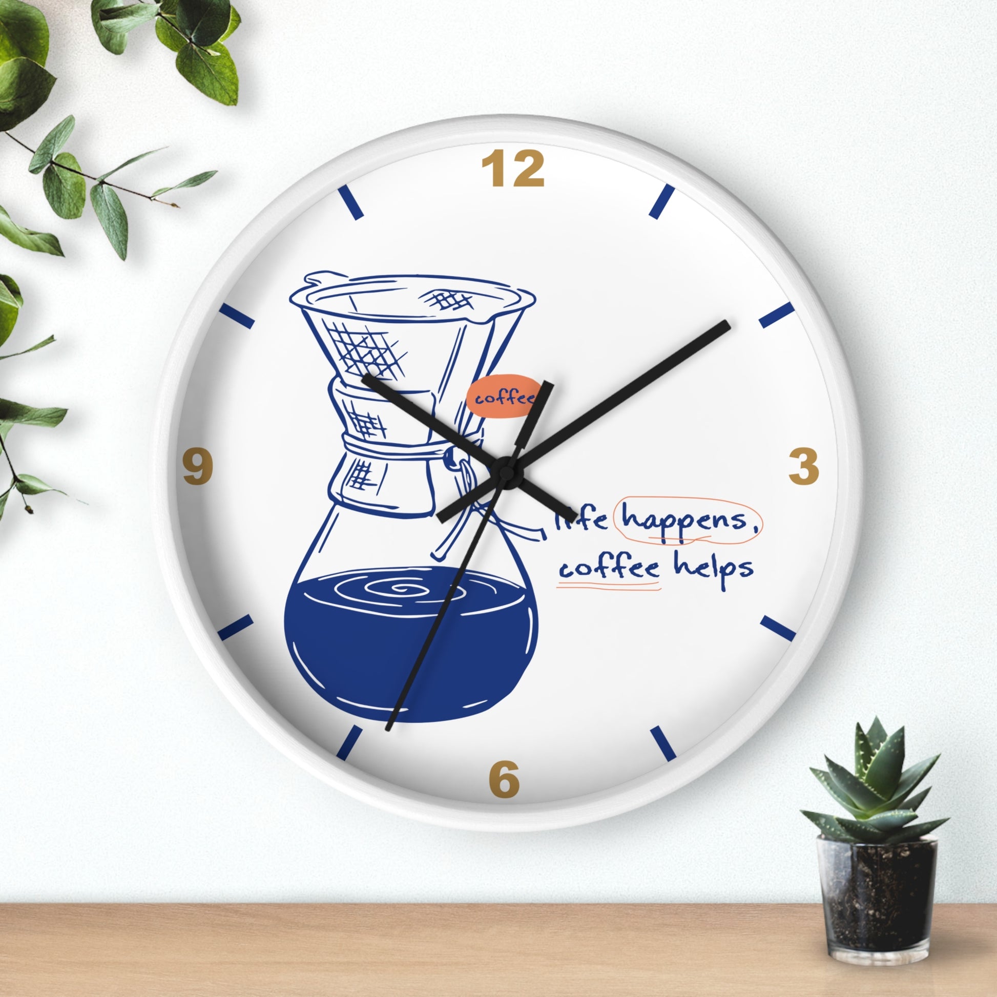 A round white wall clock with a coffee-themed design, featuring a chafing dish and pour-over coffee illustration, blue hands, gold numbers, and the phrase 'life happens, coffee helps' on a light wall above a wooden surface with small plants.