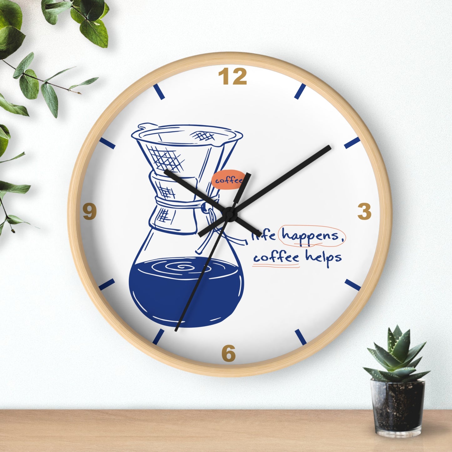 A round wall clock with a light wood frame and a white face, featuring a coffee dripper and pot illustration, blue hands, gold numerals, and the phrase 'life happens, coffee helps.' The clock is shown hanging on a white wall above a wooden surface with small green plants nearby.