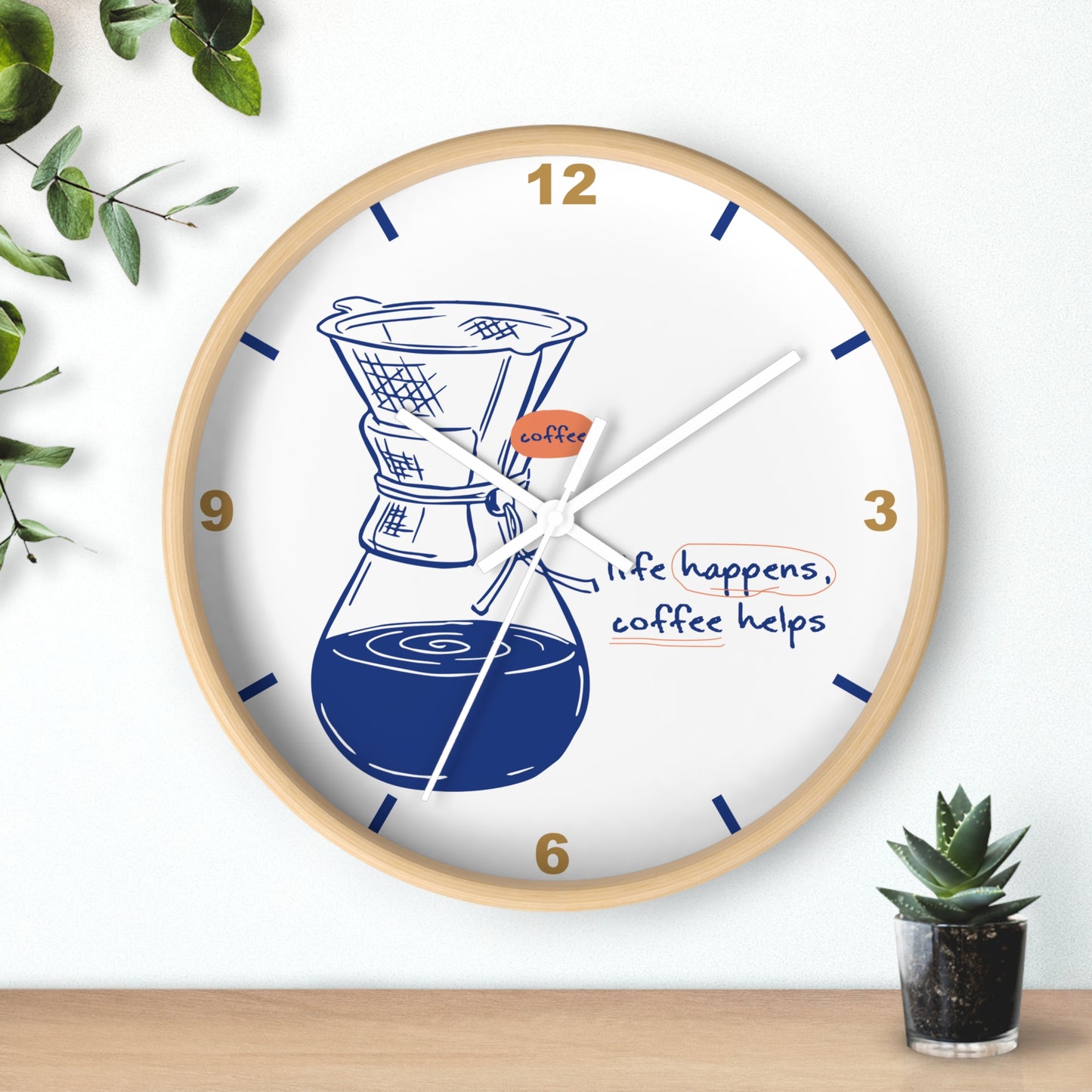 A round wall clock with a light wood frame and a white face, featuring a coffee maker illustration, gold numbers, and the phrase 'life happens, coffee helps.' The clock is shown hanging on a white wall above a wooden surface, with small green plants at the corners.