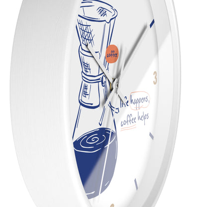 A white round wall clock with a coffee-themed design, featuring a blue line drawing of a coffee maker and cup, gold numbers, and the phrase 'life happens, coffee helps' on the dial.