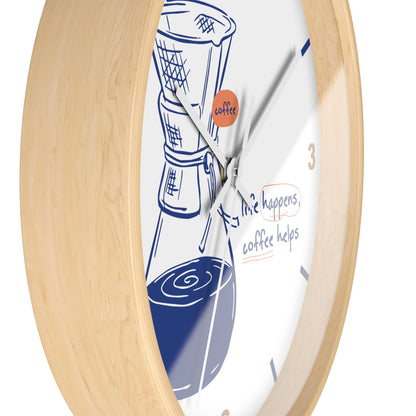 A close-up of a round wall clock with a light natural wood frame and a white face. The clock face features a blue line drawing of a coffee maker and coffee cups, with the phrase 'life happens, coffee helps' and a small orange circle containing the word 'coffee'.