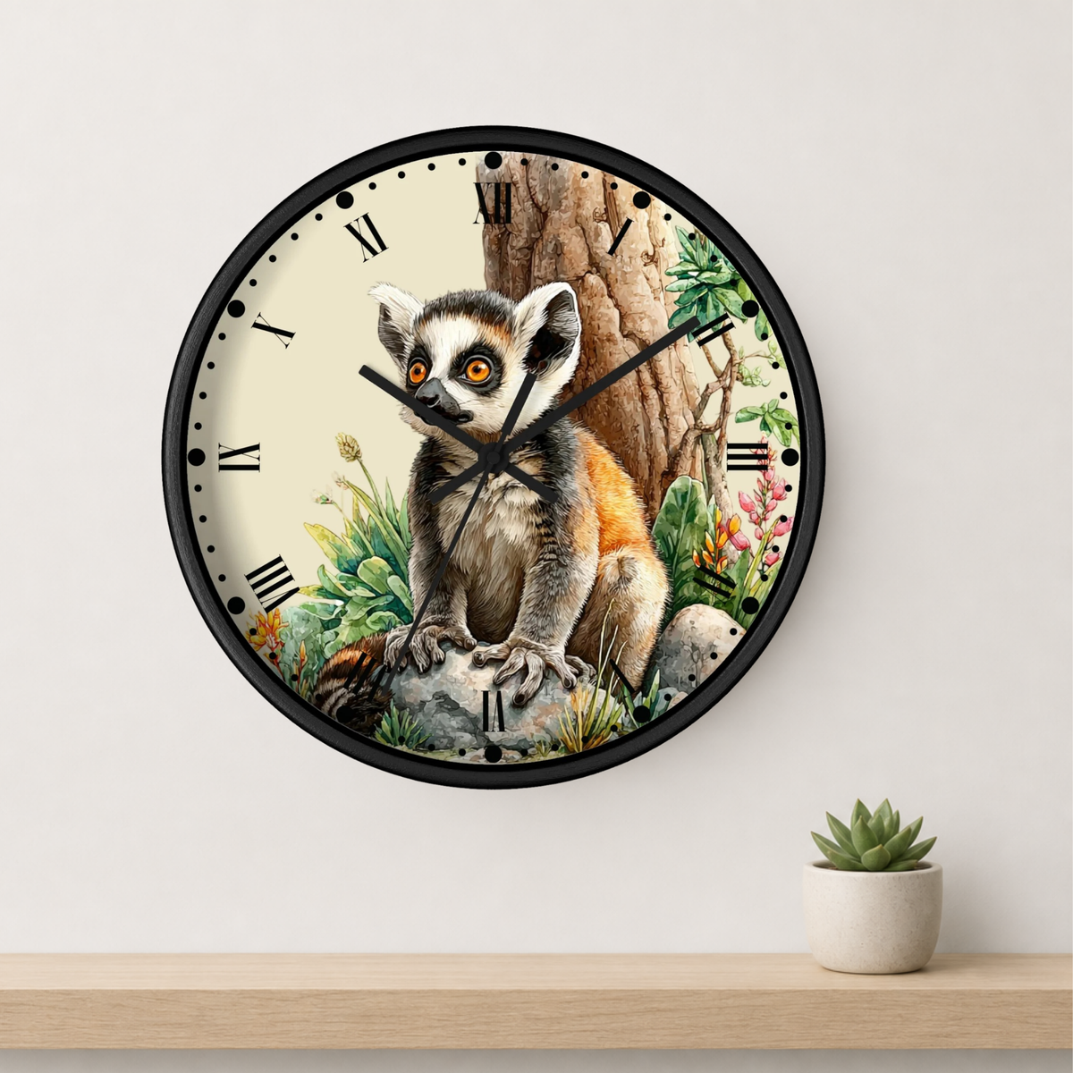 A round wall clock with a black frame and a detailed illustration of a ring-lemur sitting in front of a tree trunk, mounted on a white wall above a light wood shelf with a small potted succulent.