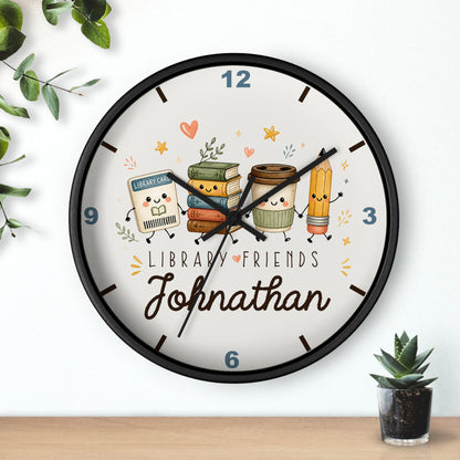 A round wall clock with a black frame and a white face, featuring cute cartoon library-themed characters and the name 'Johnathan' in script. The clock is shown hanging on a white wall above a wooden surface, with small green plants in the corners.