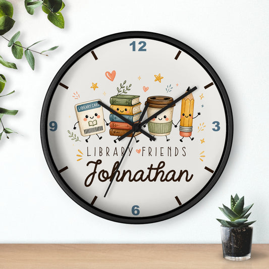 A round wall clock with a black frame and a white face, featuring cute cartoon library-themed characters and the name 'Johnathan' in script. The clock is shown hanging on a white wall above a wooden surface, with small green plants in the corners.