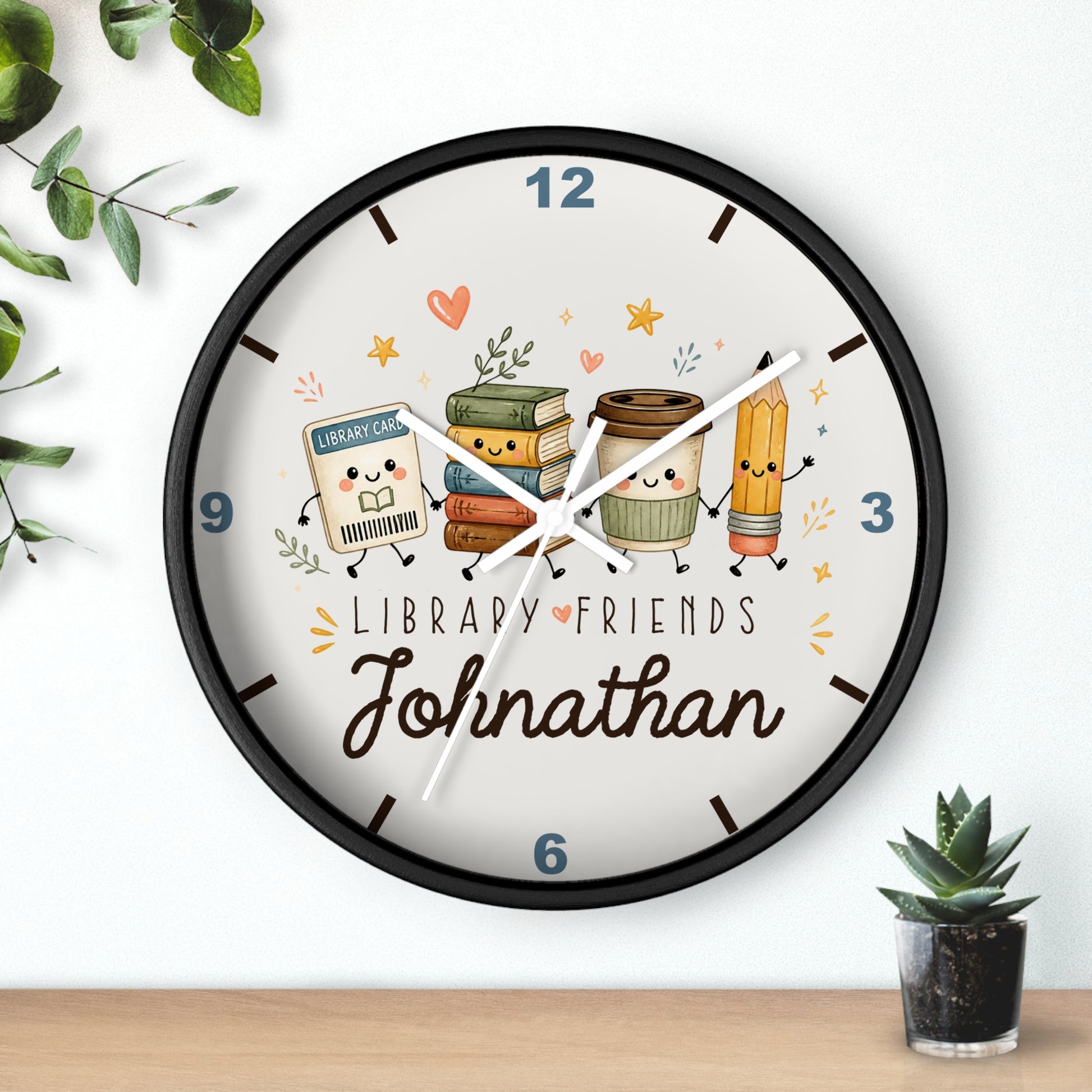 A personalized round wall clock with a black frame and a whimsical library-themed face. The clock features cute cartoon illustrations of books, a coffee cup, a pencil, and a small library card character, with the text 'LIBRARY FRIENDS' and the name 'Johnathan' in cursive.