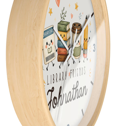 A round wall clock with a light natural wood frame and a white face featuring cute cartoon library-themed illustrations, including books, a coffee cup, a pencil, and the text 'LIBRARY FRIENDS' and 'Johnathan'.