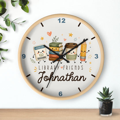 A personalized round wall clock with a light wood frame, featuring a white face decorated with cute cartoon library-themed characters and the name 'Johnathan'. The clock is styled on a wooden shelf against a white wall with decorative green leaves and a small potted succulent.