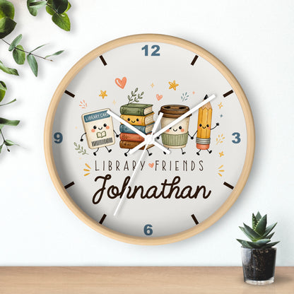 A round wooden wall clock with a light wood frame and a white face, featuring a cute illustration of library-themed characters including books, a coffee cup, and a pencil. The face is personalized with the text 'LIBRARY FRIENDS' and the name 'Johnathan' in script.