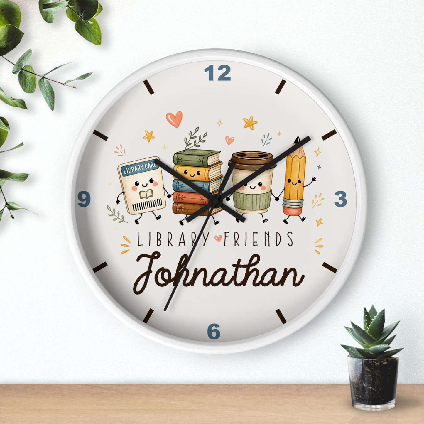 A personalized round wall clock with a white frame, featuring a cute library-themed illustration with books, a coffee cup, and a pencil. The clock face has the text 'LIBRARY FRIENDS' and the name 'Johnathan' in script.