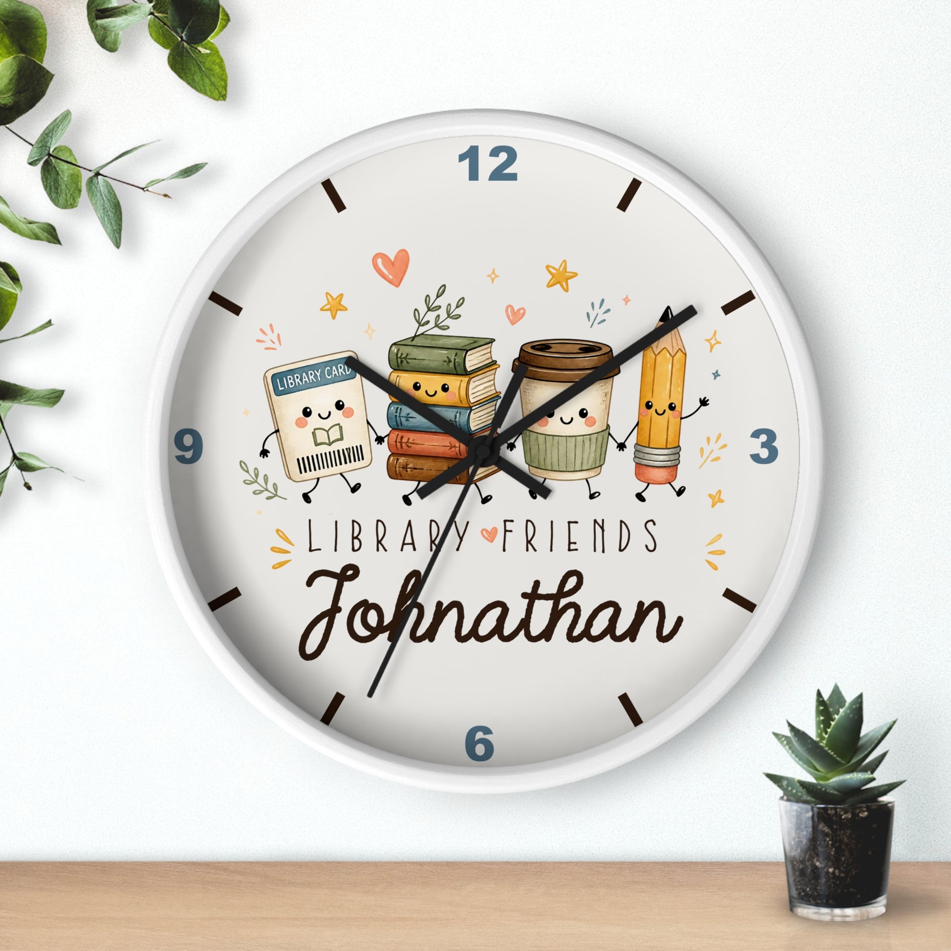 A personalized round wall clock with a white frame, featuring a cute library-themed illustration with books, a coffee cup, and a pencil. The clock face has the text 'LIBRARY FRIENDS' and the name 'Johnathan' in script.
