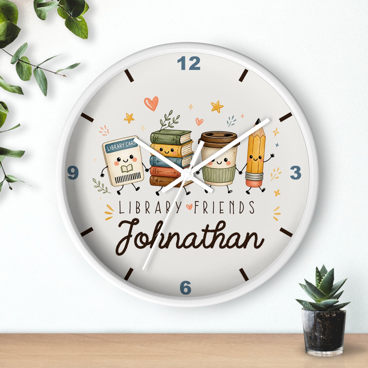 A personalized round wall clock with a white frame, featuring cute illustrated library-themed characters and the name 'Johnathan' on the clock face, styled on a light wall above a wooden surface with small plants.