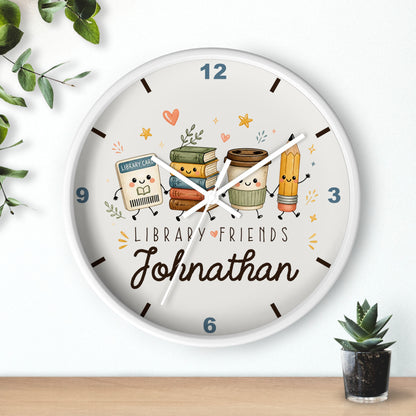 A personalized round wall clock with a white frame, featuring cute illustrated library-themed characters and the name 'Johnathan' on the clock face, styled on a light wall above a wooden surface with small plants.
