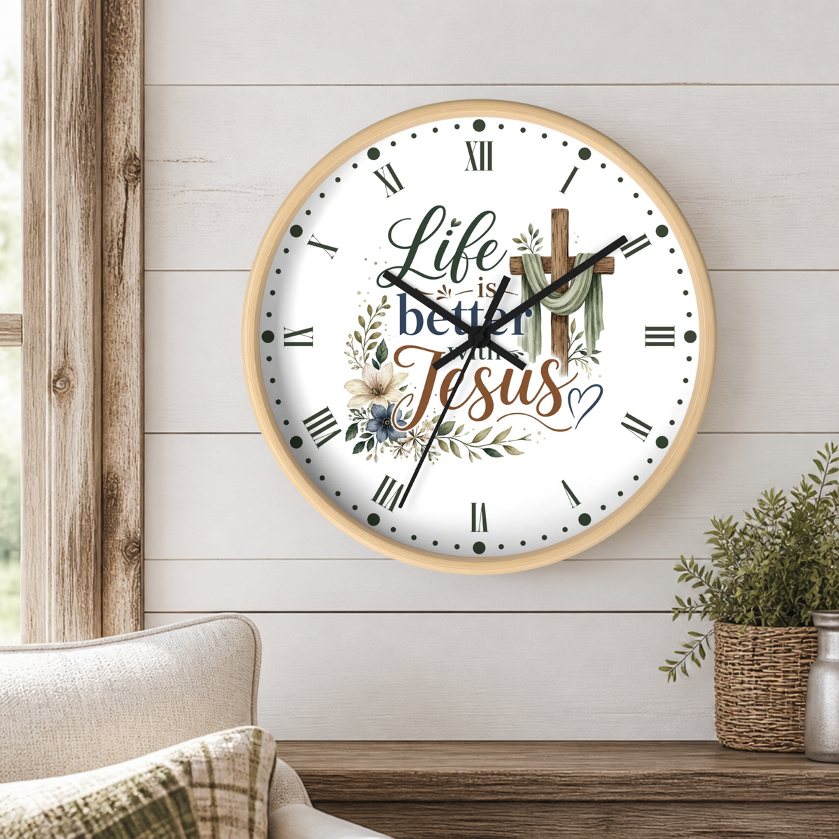 A round wall clock with a light wood frame hangs on a white shiplap wall above a wooden table. The clock face is white with black Roman numerals, black hands, and the phrase 'Life is better with Jesus' in decorative script, decorated with floral and cross illustrations.