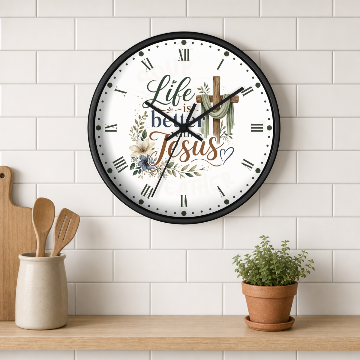 A round wall clock with a black frame and a white face featuring the text 'Life is better  with Jesus' with floral and cross illustrations, mounted on a white subway tile wall above a wooden shelf with kitchen utensils and a potted plant.