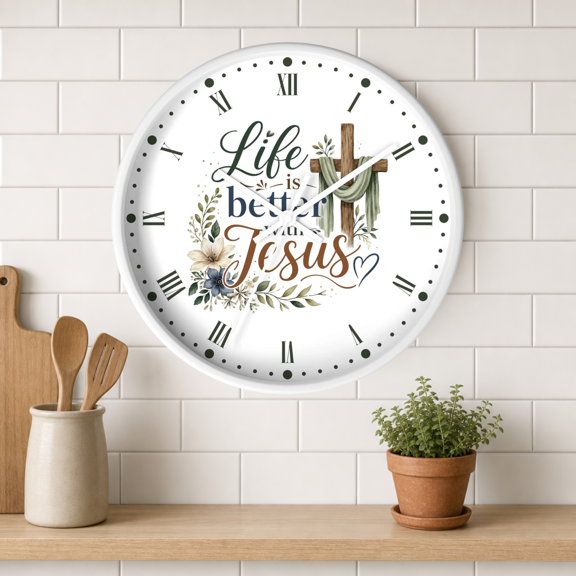 A round white wall clock with Roman numerals and the phrase 'Life is better with Jesus' in decorative script and floral artwork, mounted on a white subway tile wall above a wooden shelf with kitchen utensils and a potted plant.