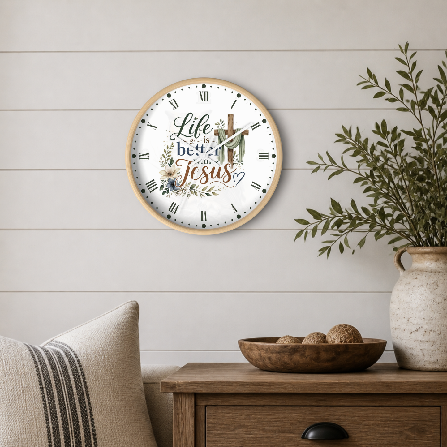 A round wall clock with a light wood frame and a white face featuring the phrase 'Life is better with Jesus' in script and serif fonts, surrounded by floral and cross illustrations. The clock is mounted on a white shiplap wall above a wooden side table with a bowl, decorative pillows, and a potted plant.