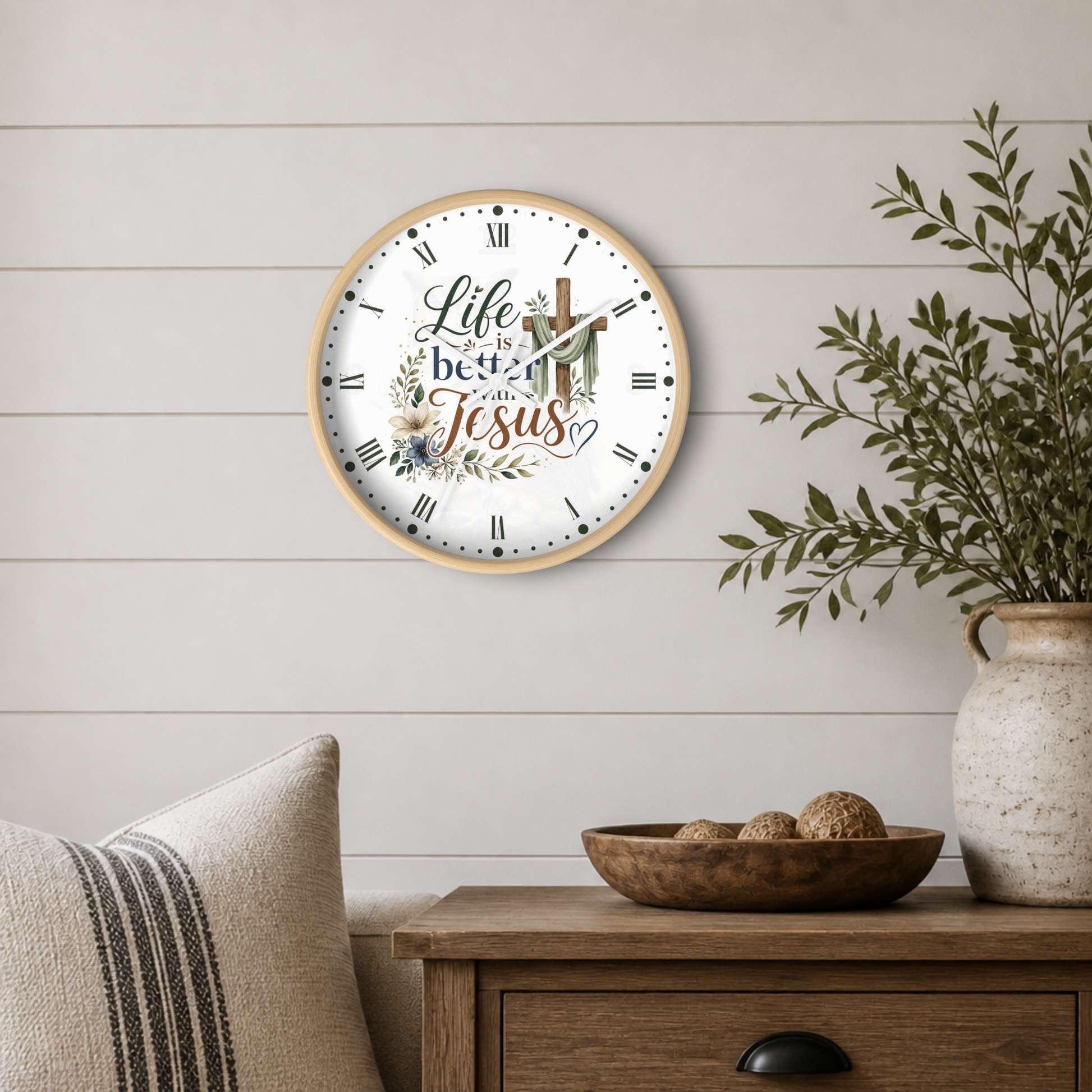 A round wall clock with a light wood frame and a white face featuring the phrase 'Life is better with Jesus' in script and serif fonts, surrounded by floral and cross illustrations. The clock is mounted on a white shiplap wall above a wooden side table with a bowl, decorative pillows, and a potted plant.
