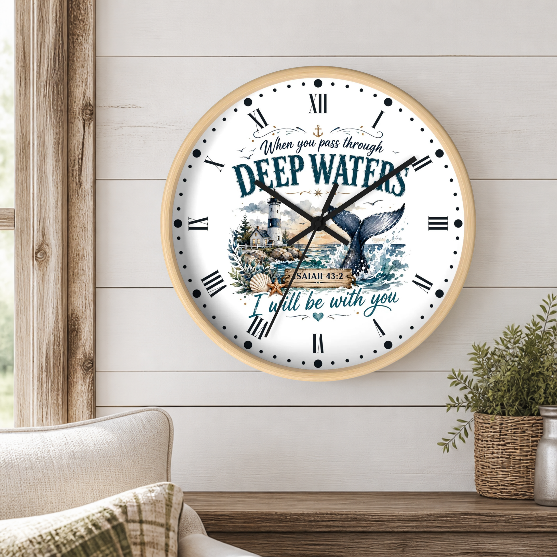 A round wall clock with a light wood frame hangs on a white shiplap wall above a wooden table. The clock face is white with black Roman numerals, black hands, and a central illustration of a lighthouse and a whale tail, surrounded by the words 'When you pass through DEEP WATERS I will be with you' and 'ISAAC 42:5'.