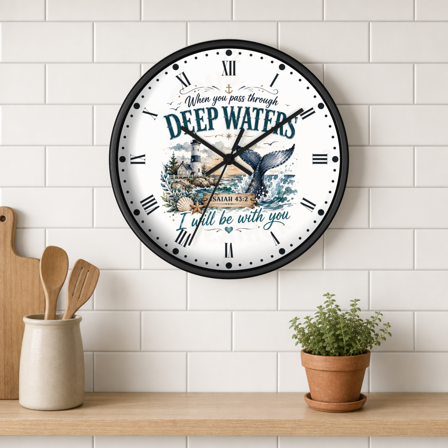 A round wall clock with a black frame hangs on a white subway tile wall above a wooden shelf. The clock face is white with a black Roman numeral hour markers and a central illustration featuring a whale tail, a lighthouse, and the text 'When you pass through DEEP WATERS I will be with you' and 'ISAIAH 42:2'.