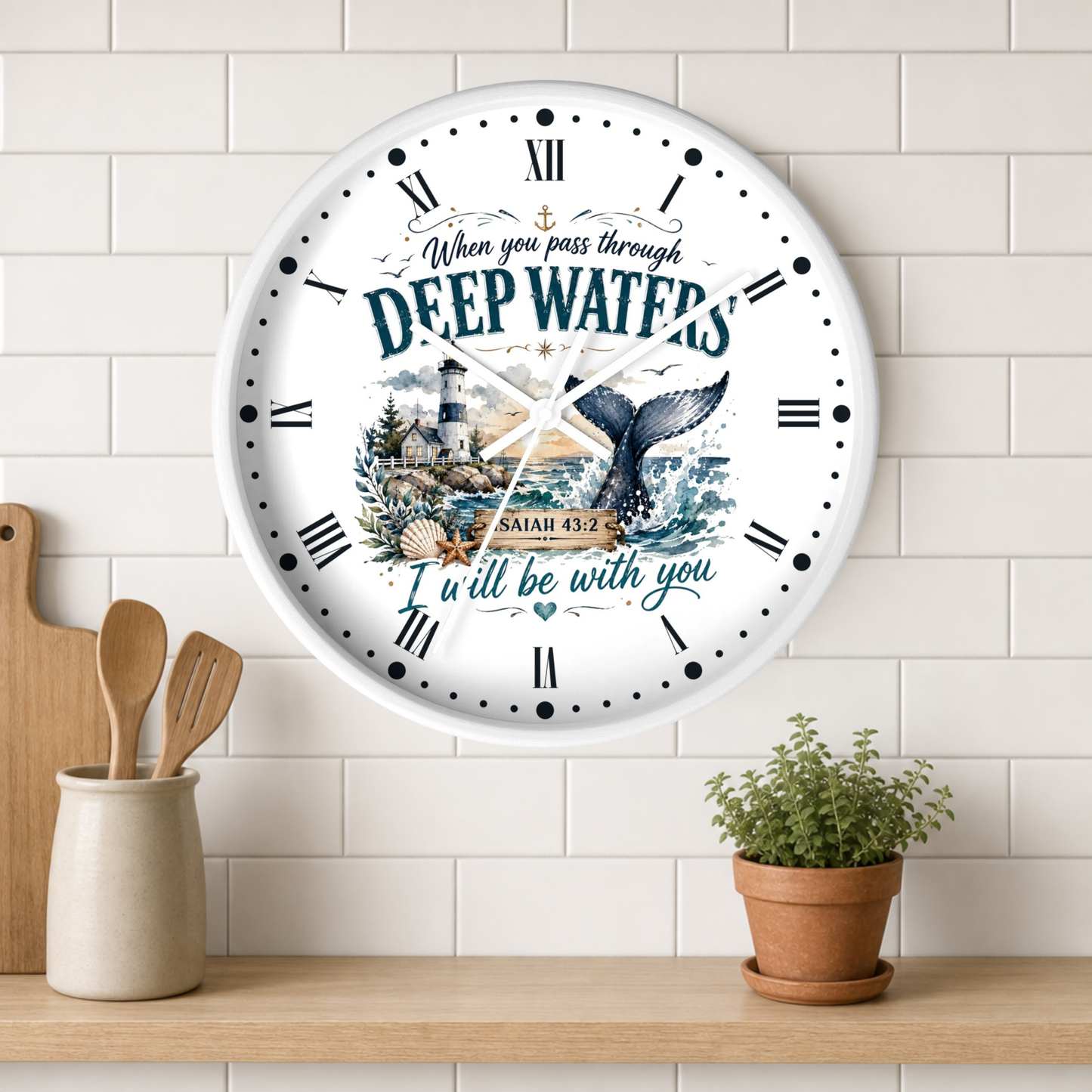A round white wall clock with Roman numerals and a nautical illustration featuring a lighthouse, sea turtle, and the words 'DEEP WATERS' and 'ISAIAH 43:2', shown hanging on a white subway tile wall above a wooden shelf with a small potted plant and utensils.