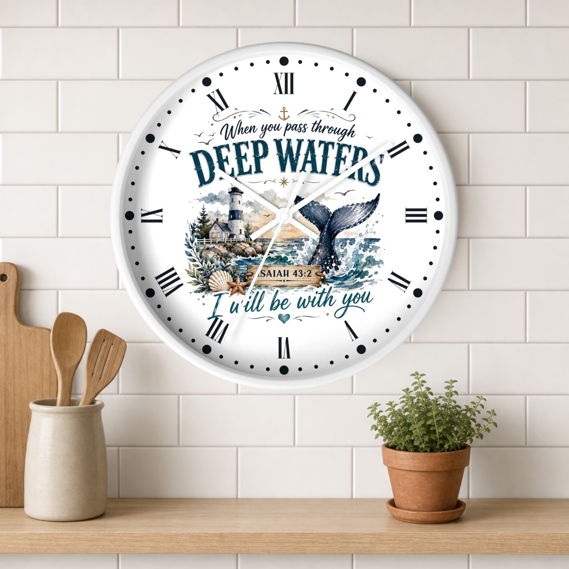 A round white wall clock with Roman numerals and a nautical illustration featuring a lighthouse, sea turtle, and the words 'DEEP WATERS' and 'ISAIAH 43:2', shown hanging on a white subway tile wall above a wooden shelf with a small potted plant and utensils.