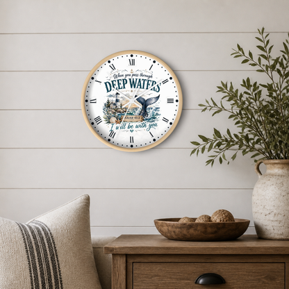 A round wall clock with a light wood frame hangs on a white shiplap wall above a wooden side table. The clock face is white with navy blue Roman numerals, a nautical illustration featuring a whale tail and ship, and the text 'When you leave through DEEP WATERS I'll be with you.