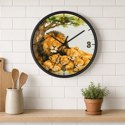 A round wall clock with a black frame and a watercolor-style lion family illustration, mounted on a white subway tile wall above a wooden shelf with kitchen utensils and a potted plant.