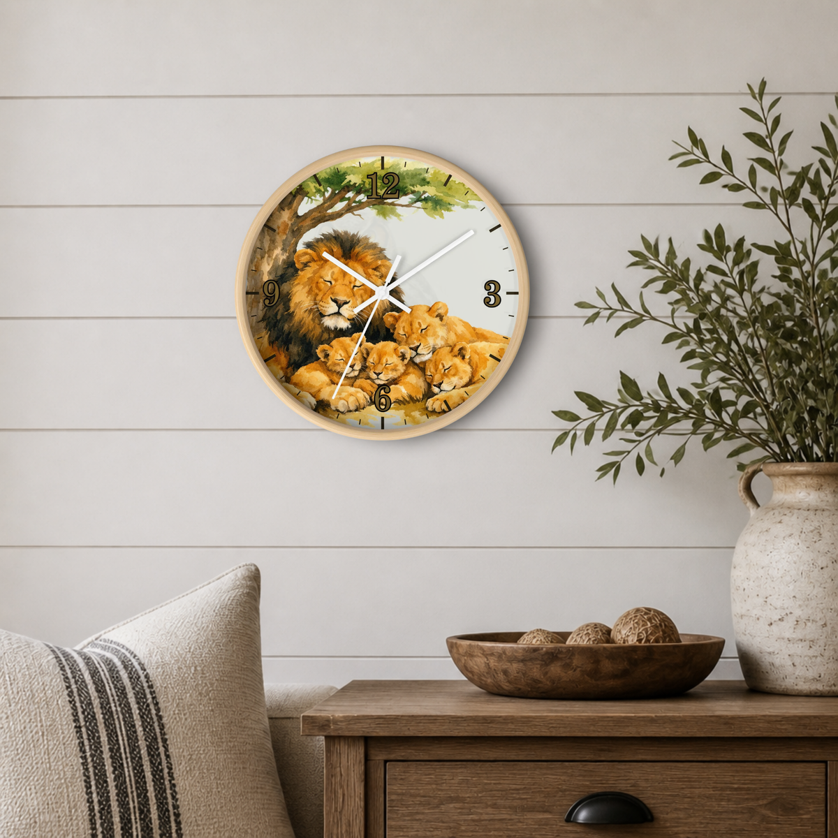 A round wall clock featuring a painted lion family scene, mounted on a white shiplap wall above a wooden side table with a vase and bowl.