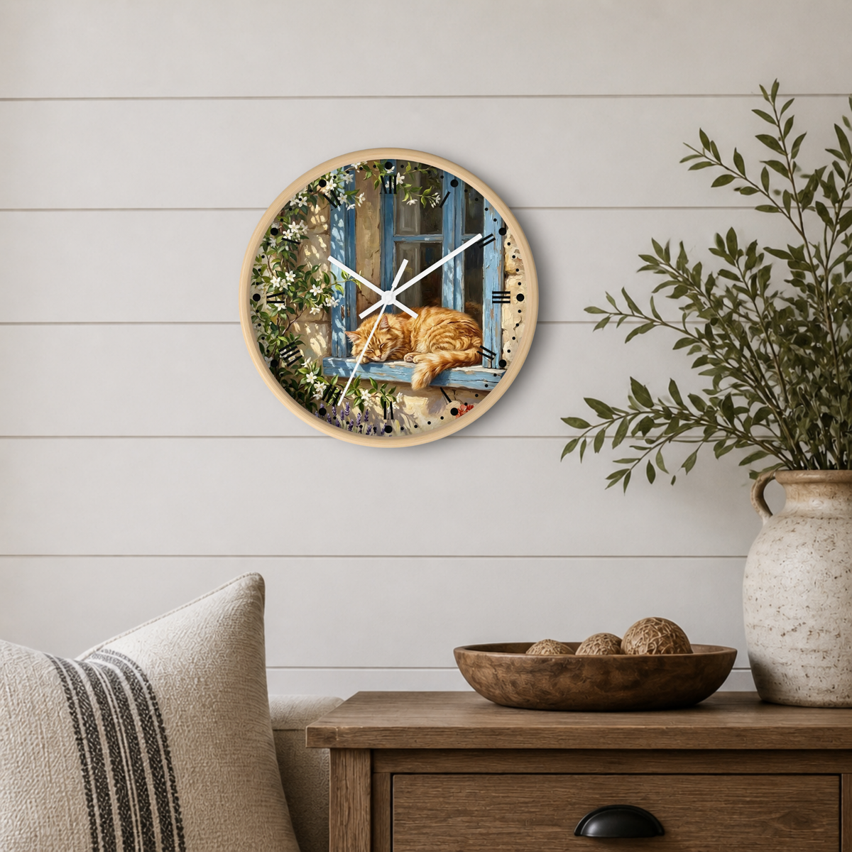 Round wall clock with a light wood frame featuring a printed illustration of an orange tabby cat sleeping on a blue window sill, mounted on a white shiplap wall above a wooden side table with a plant and decorative bowl.