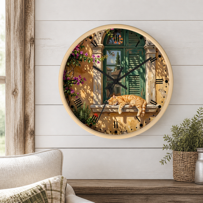 A round wall clock with a light wood frame hangs on a white shiplap wall above a wooden table. The clock face features a detailed illustration of an orange tabby cat sleeping on a windowsill of a Mediterranean-style building with a green window and purple flowers.