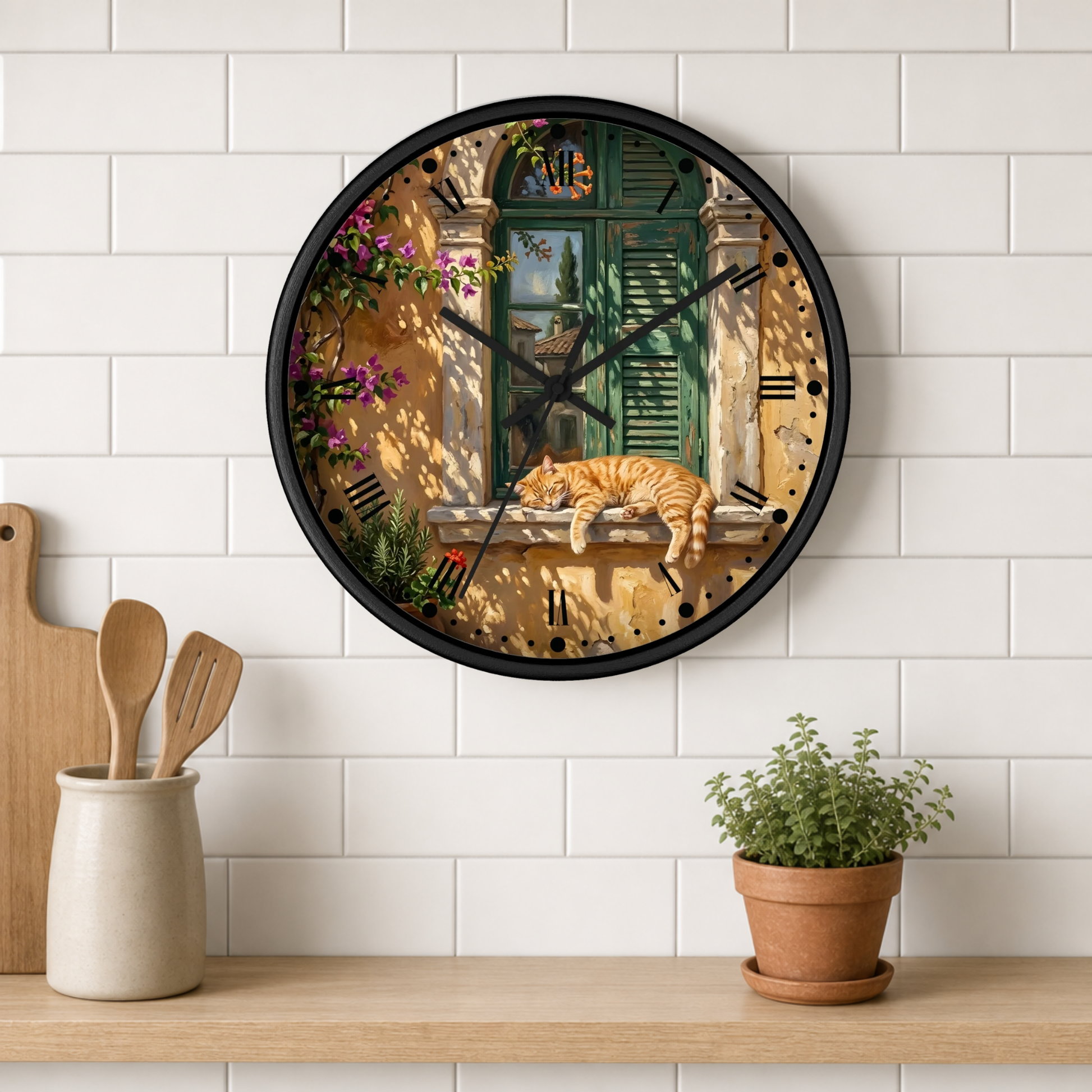 A round wall clock with a black frame hangs on a white subway-tile kitchen wall above a wooden shelf. The clock face features a detailed illustration of an orange tabby cat sleeping on a windowsill in front of green shutters, with pink flowers and a warm yellow-beige background.