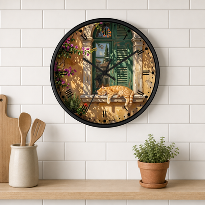 A round wall clock with a black frame hangs on a white subway-tile kitchen wall above a wooden shelf. The clock face features a detailed illustration of an orange tabby cat sleeping on a windowsill in front of green shutters, with pink flowers and a warm yellow-beige background.