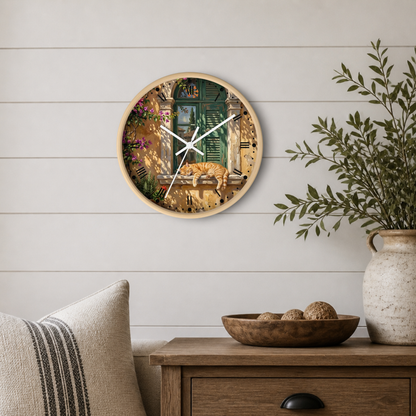 Round wall clock with a light wood frame and a scenic print of a rustic courtyard scene of an orange tabby cat sleeping in a window , styled on a white shiplap wall above a wooden side table with a plant and decorative bowl.