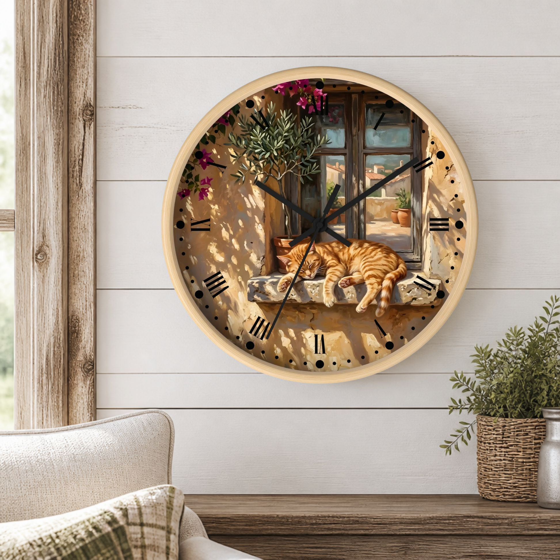 A round wall clock with a light wood frame hangs on a white shiplap wall above a wooden table. The clock face features a scenic illustration of an orange tabby cat sleeping on a windowsill next to a window with hanging green plants and pink flowers.