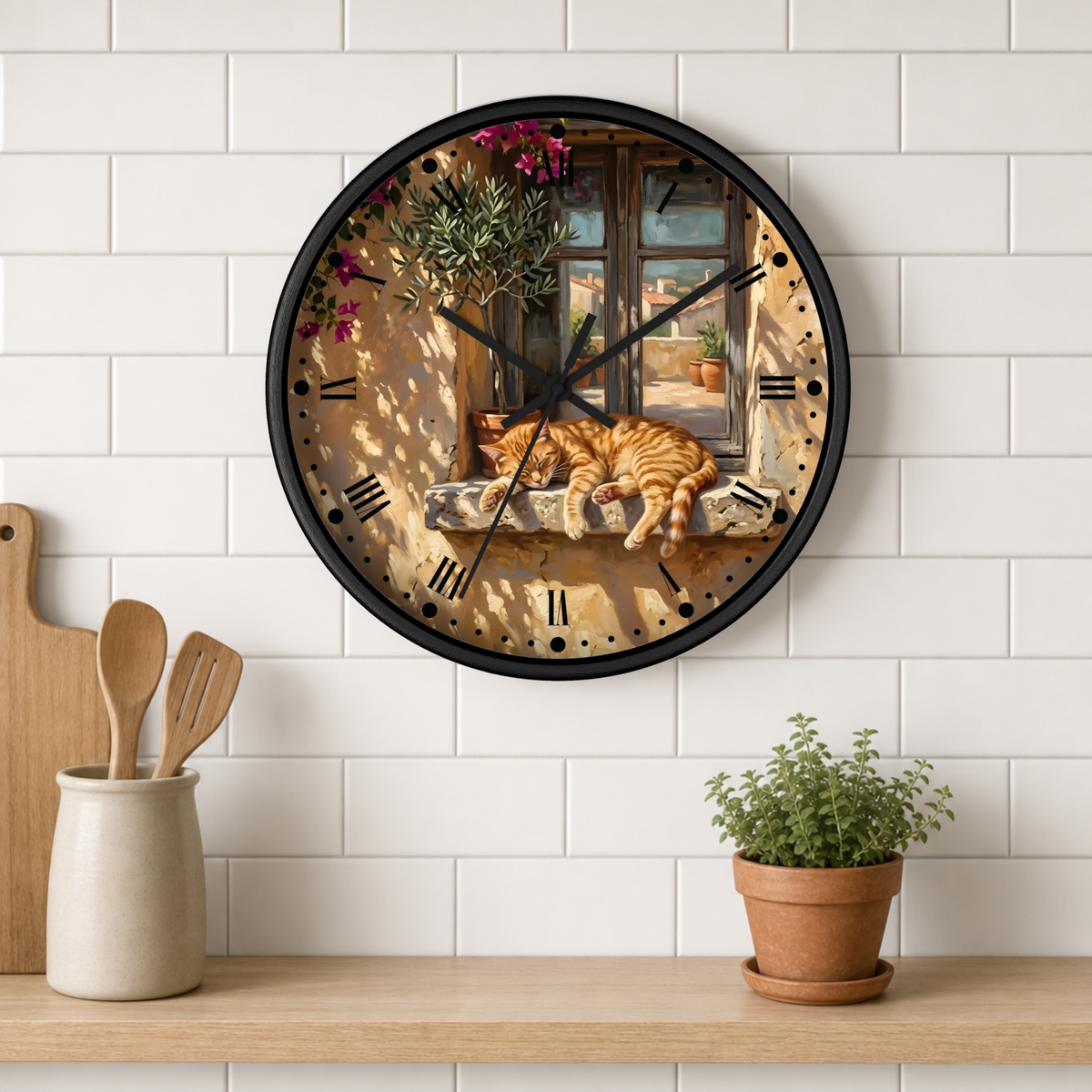 A round wall clock with a black frame and a printed scene of an orange tabby cat sleeping by a window, mounted on a white subway tile wall above a wooden shelf with kitchen utensils and a potted plant.
