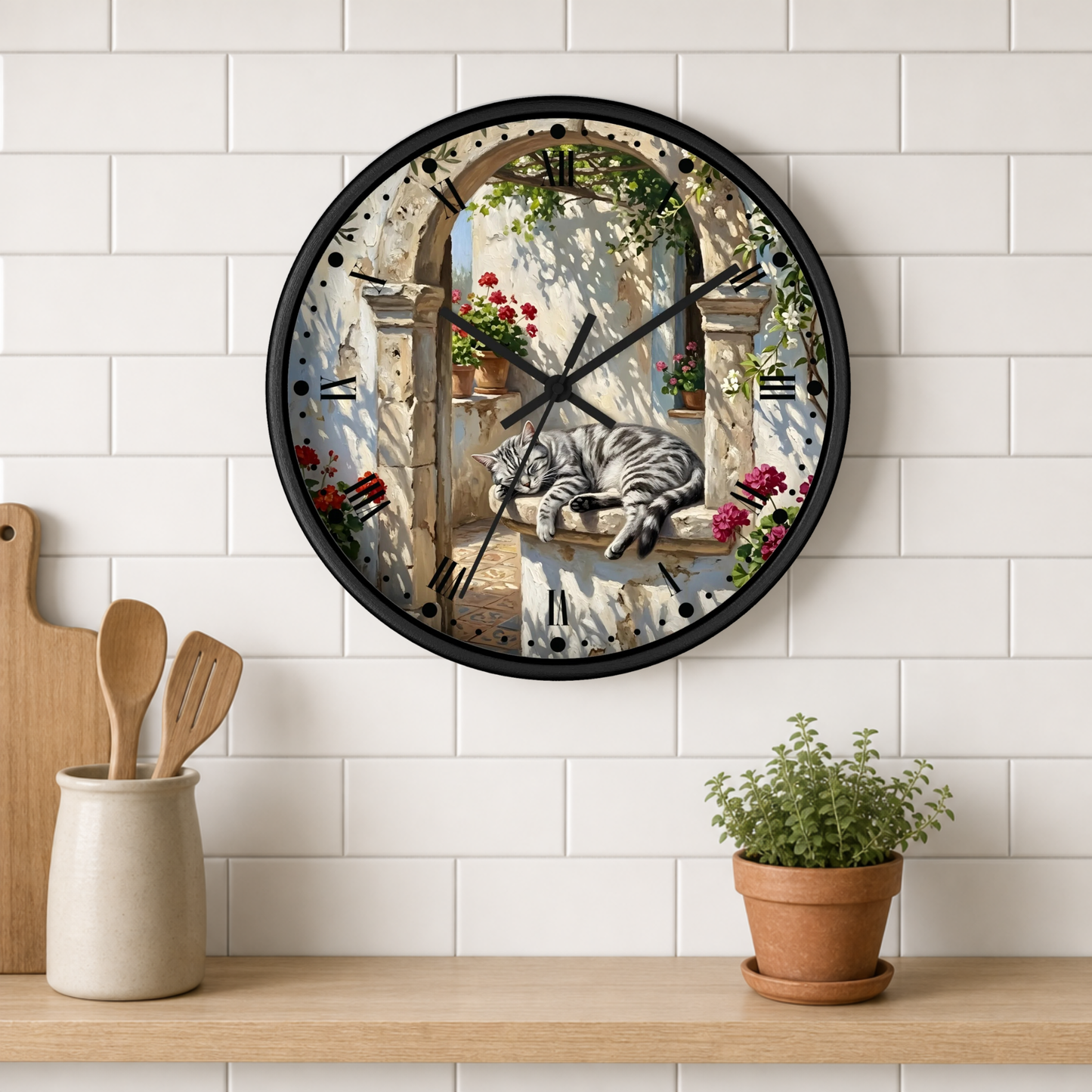 A round wall clock with a black frame hangs on a white subway-tile kitchen wall above a wooden shelf. The clock face features a printed illustration of a gray tabby cat sleeping on a stone ledge in front of an arched doorway with red flowers.