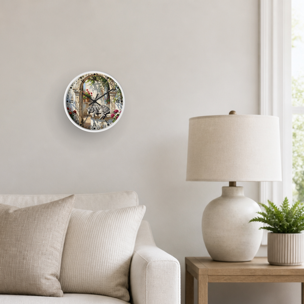 A round wall clock with a detailed grey tabby cat sleeping illustration hangs on a light beige wall in a styled living room setting next to a table lamp and sofa