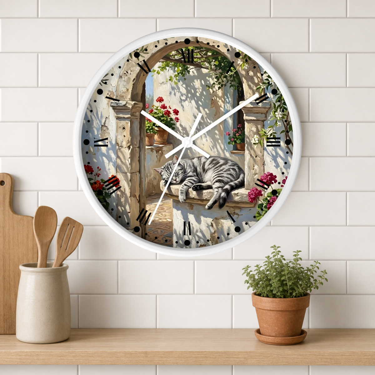 A round wall clock with a white frame hangs on a white subway-tile wall above a wooden shelf. The clock face features a scenic illustration of a stone archway with flowers, greenery, and a sleeping gray tabby cat.