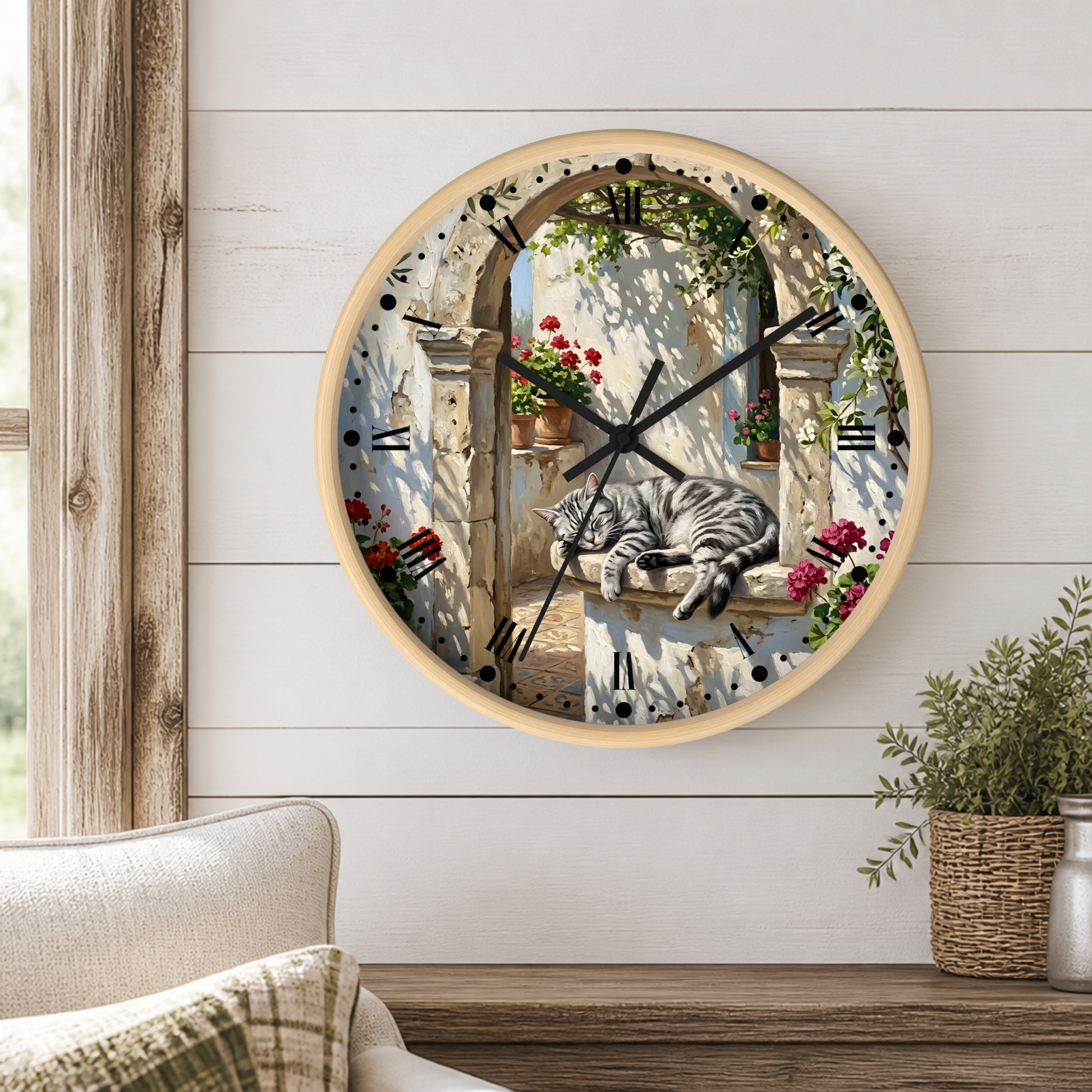 A round wall clock with a light wood frame hangs on a white shiplap wall above a wooden table. The clock face features a scenic illustration of a gray tabby cat sleeping on a stone windowsill with red flowers and green vines, set against a blue and beige background.