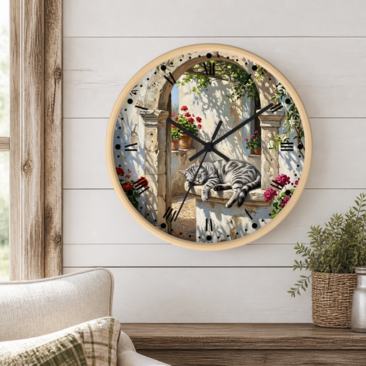 A round wall clock with a light wood frame hangs on a white shiplap wall above a wooden table. The clock face features a scenic illustration of a gray tabby cat sleeping on a stone windowsill with red flowers and green vines, set against a blue and beige background.