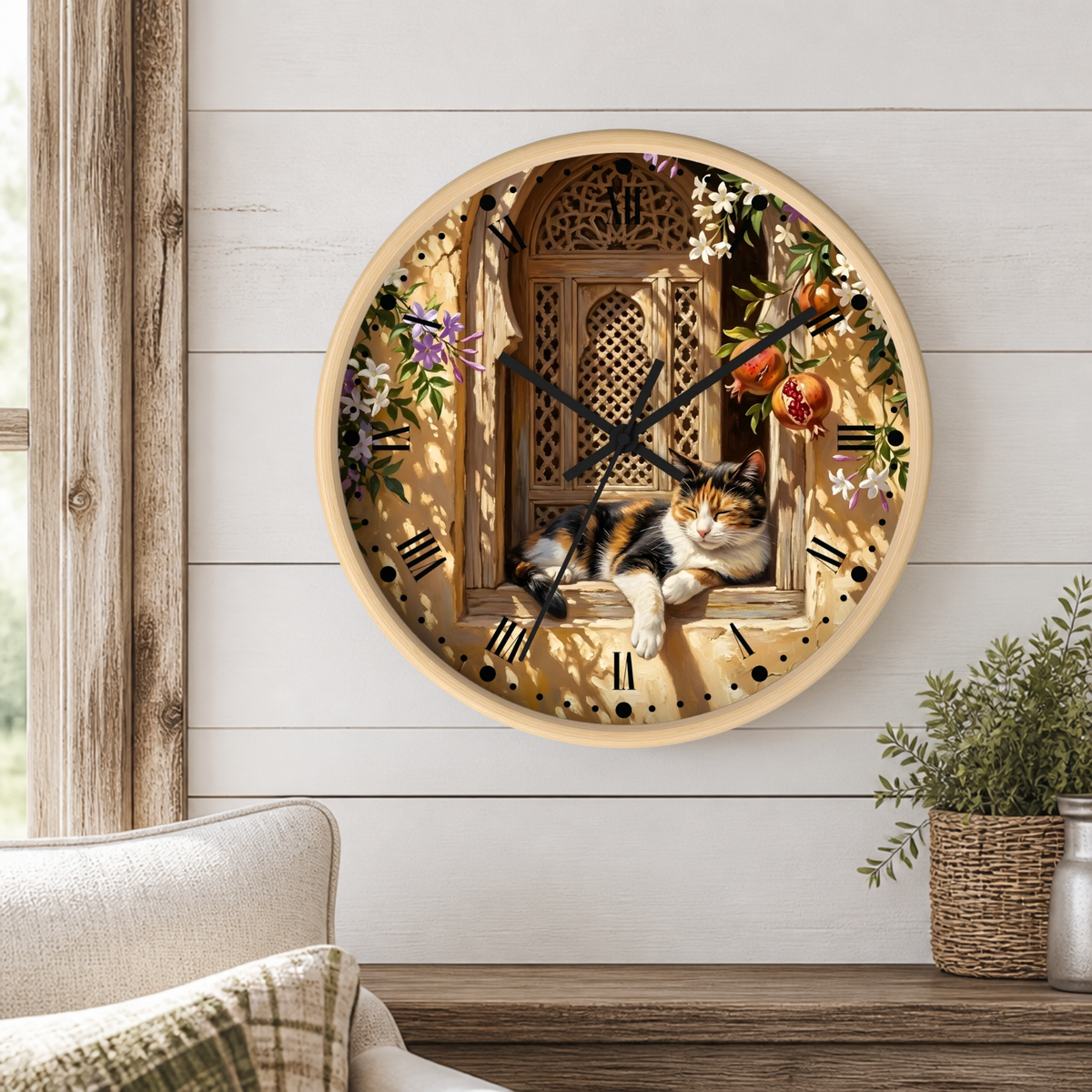 A round wall clock with a light wood frame hangs on a white shiplap wall above a wooden table. The clock face features a detailed illustration of a calico cat lying on a windowsill with a wooden window pane behind it, surrounded by flowers and fruit.