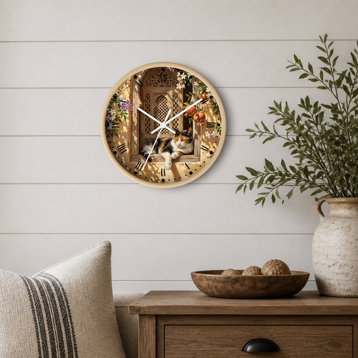 Round wall clock with a light wood frame and a printed scene of a cat sleeping in a window, styled on a white shiplap wall above a wooden side table with a plant and decorative bowl