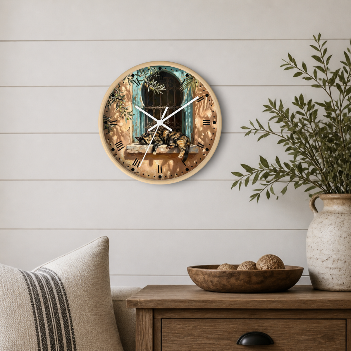 A round wall clock with a rustic illustration of a cat on a windowsill, styled on a white shiplap wall above a wooden side table with a plant and decorative bowl.